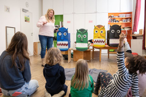 Vor einer Kindergruppe sind auf vier Stühlen vier bunte Monsterchen platziert, durch dessen Münder man etwas einwerfen kann.