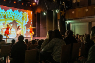 Es ist eine Bühne zu sehen, auf der Ballett getanzt wird, sowie ein Teil des Publikums von hinten. In der Mitte des Bildes sitzt ein Mädchen auf dem Schoß einer Frau, beide schauen zur Bühne.