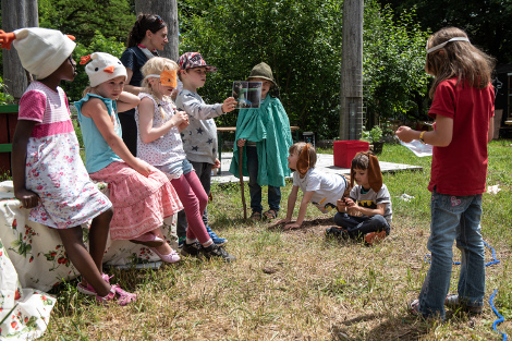 Mehrere Kinder mit Masken und Verkleidungen sitzen und stehen auf einer Wiese bei gutem Wetter