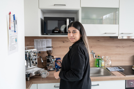 Ein Frau mit langen schwarzen Haaren steht vor einer Kaffeemaschine und hält einen Siebträger in der hand