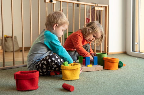 Zwei Kleinkinder sind in ein Holzspiel versunken, bei dem Klötze nach ihrer Form einsortiert werden.