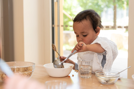 Ein Kleinkind sitzt mit Lätzchen am Tisch und isst selbst mit einem Löffel aus einer Schüssel