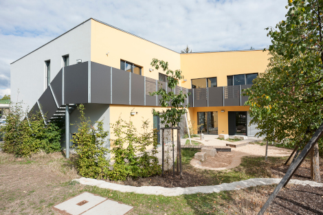 Bild des Gebäudes von außen: ein gelbgestrichener Neubau mit Grünfläche drumherum.