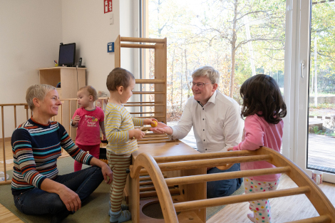 Ein Mann in hellem Oberhemd sitzt mit ein paar Kindern und einer Erzieherin am Boden und reicht lachend einem Kind ein Spielzeug.