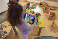 Ein M&auml;dchen, von hinten zu sehen, steht in einem Gruppenraum der Kita, h&auml;lt ein Tablet in der Hand und schaut darauf hinab.