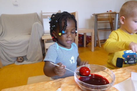 Ein lachendes Kind sitzt am Tisch und hält ein Ei auf einem Löffel in eine Schale mit roter Farbe.