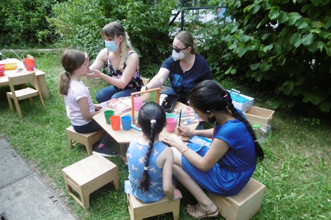 An einem Tisch im Garten sitzen mehrere Kinder, die von zwei Frauen geschminkt werden