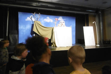 Vorne sind ein paar Hinterköpfe von Kindern, im Hintergrund eine Bühne mit einigen Requisiten und Bühnenbild