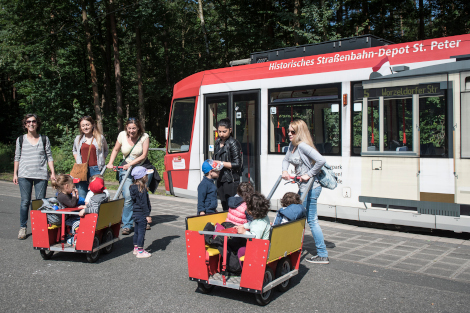 Erzieherinnen stehen an einer Tramhaltestelle mit zwei Bollerwagen, in denen Kinder sitzen.