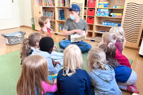 Eine Gruppe Kinder sitzt mit dem Rücken zur Kamera im Halbkreis, vor ihnen ein junger Mann, der ein Bilderbuch hält und erzählt.