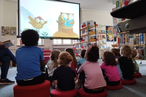 Kinder sitzen auf roten Sitzkissen am Boden, vor Ihnen ist eine Leinwand mit einem Bild von zwei Eulen