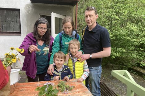 Zwei Kinder und drei Erwachsene stehen in einem Garten vor einem Tisch, auf dem ein Haufen Kräuter liegen.