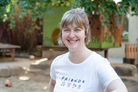 Eine Frau mit kinnlangen blonden Haaren und weißem Shirt steht draußen im Innenhof von MOMO und lacht in die Kamera. 