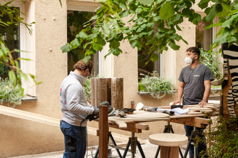 Zwei junge Männer stehen draußen in einem Hof mit Werkzeugen an einem Tisch und bearbeiten Holz.