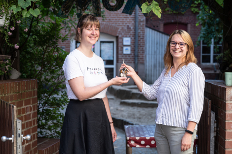 Zwei Frauen stehen in einem Innenhof. Die eine reicht der anderen ein Schlüsselbund