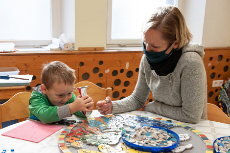 Eine Frau mit Alltagsmaske und ein Junge sitzen an einem Tisch und kleben gemeinsam bunte Scherben auf ein großes Mosaik.