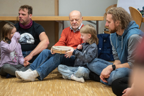 Kinder sitzen mit ihren Vätern am Boden und probieren Percussion-Instrumente aus.