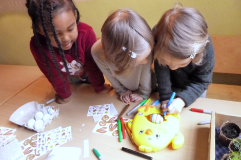 Drei Kinder beugen sich über einen Tisch mit Bastelmaterialien und eine Eierdrehmaschine, in der sie ein Ei bemalen.