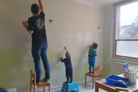 Ein Mann und zwei Kinder stehen auf Stühlen mit Pinseln in der Hand und streichen eine Wand.