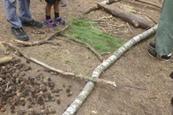Auf einem Waldboden ist mit unterschiedlichen Materialien ein Barfußpfad gelegt. Drum herum stehen Kinder und eine Erwachsene, wobei nur die Beine knieabwärts zu sehen sind.