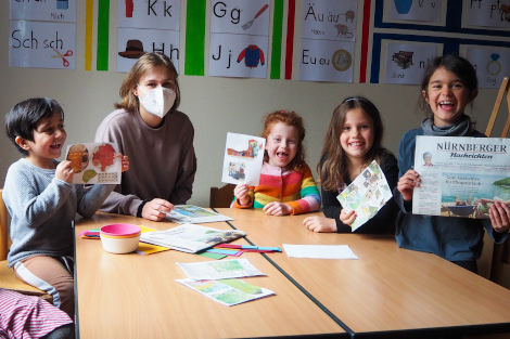 Am Tisch sitzt eine Frau mit mehreren Kindern, die stolz Zeitungen und Zeitungscollagen hochhalten.