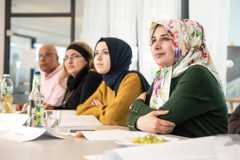 Mehrere Frauen sitzen in einer Infoveranstaltung und hören interessiert zu. Zeit von ihnen tragen ein Kopftuch.