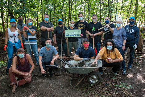 15 Menschen stehen in einem Waldstück als Gruppe zusammen mit Masken auf den Gesichtern und Gartenwerkzeuge in den Händen. Im Vorderung steht ein mit Steinen beladener Schuppkarren.