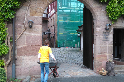 Eine Frau mit einem gelben Dolby-T-Shirt schiebt eine Schubkarre durch eine großes Sandsteintor