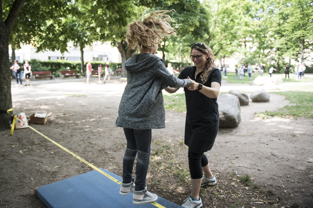 Ein ca. 12-jähriges Mädchen steht auf einer Slackline, die langen Haare schwingen in der Luft. Sie wird von einer Frau mit schwarzer Kleidung an den Händen gehalten.