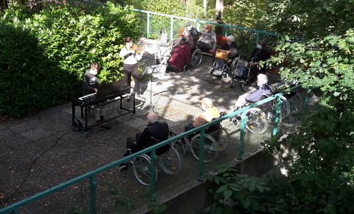 Zwei mehrstöckige Häuser durch einen Garten und eine Terrasse verbunden, in dem ältere Menschen z.T. in Rollstühlen sitzen und einem Musiker am Klavier zuhören.