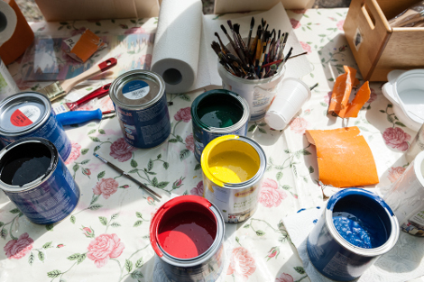 Auf einem Tisch mit Tischdecke stehen offene Lackdosen mit bunten Farben und ein Becher mit Pinseln