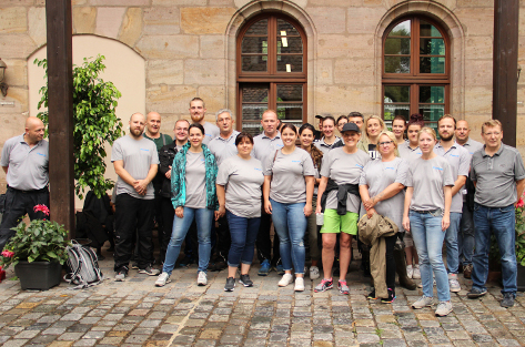 20 Personen, die meisten mit grauen T-Shirts mit der Aufschrift Fürst, stehen vor einem Haus und schauen in die Kamera.