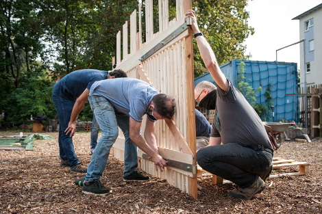 Drei Männer im Freien arbeiten an einer Holzwand, die sie von links und rechts stützen. 