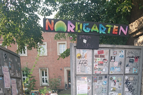 Buntes Schild "Nordgarten" hängt über dem Eingangstor zu einem großen Garten. Dort ist im Hintergrund ein Ziegelstein-Haus zu sehen.