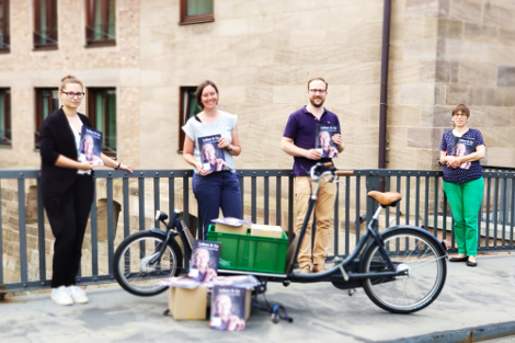 An einem Geländer stehen drei Frauen und ein Mann, diese halten Zeitschriften in der Hand. Vor ihnen steht ein Lastenfahrrad in dem weitere Kartons mit Zeitschriften zu sehen sind.