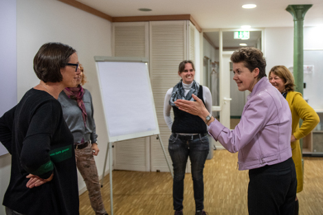 Moderation fünf Frauein Stehen im Kreis. Eine Frau mit kurzen Haaren udn einer rosa Jacke weist mit ihrer Hand zu ihrer Nachbarin auf der linken Seite.