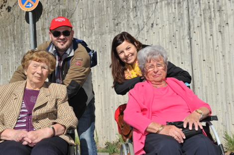 Zwei ältere Damen in Rollstühlen lachen gemeinsam mit einem Mann und einer Frau von Novartis in die Kamera.