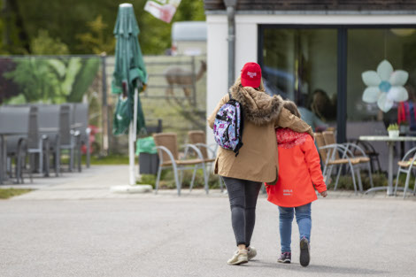 Eine Frau in brauner Jacke und ein Mädchen in roter Jacke von hinten. Die Frau legt ihren Arm um die Schulter des Kindes.