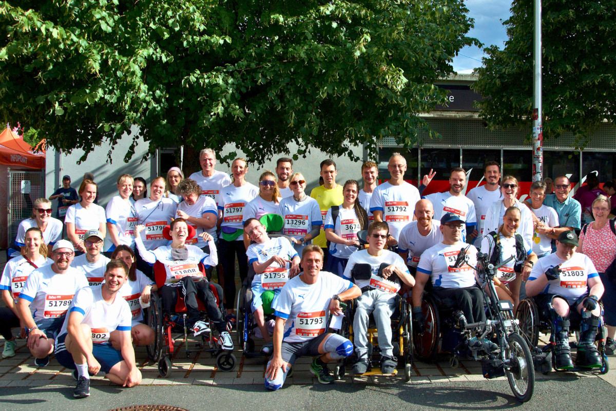 Dreißig Personen stehen bei einer Laufveranstaltung für ein Gruppenbild zusammen und haben weiße T-Shirts an. In der vorderen Reihe sitzen einige Personen im Rollstuhl.