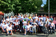 Dreißig Personen stehen bei einer Laufveranstaltung für ein Gruppenbild zusammen und haben weiße T-Shirts an. In der vorderen Reihe sitzen einige Personen im Rollstuhl.