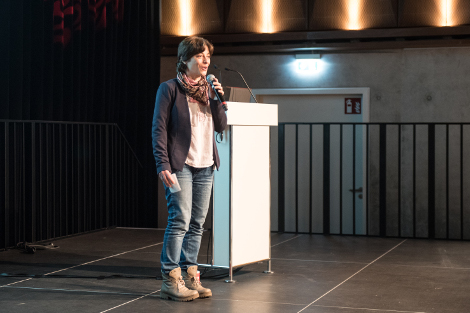 Auf einer leeren Bühne steht eine Frau mit Jeans, Strickjacke und kurzen dunklen Haaren. Sie spricht fröhlich lächelnd in ein Mikrofon.