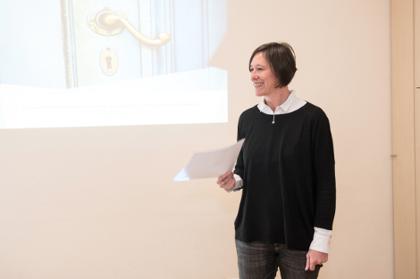 Eine Frau mit halblangen, dunklen Haaren und einem schwarzen Pulover steht mit Zetteln in der Hand vor einer Präsentation