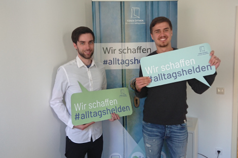 Vor einem Rollup mit einer blauen Tür stehen zwei junge Männner und lächeln in die Kamera. Sie halten Sprechblasen aus Papier in der Hand auf denen steht: "Türen Öffnen - Wir schaffen #Alltagshelden"