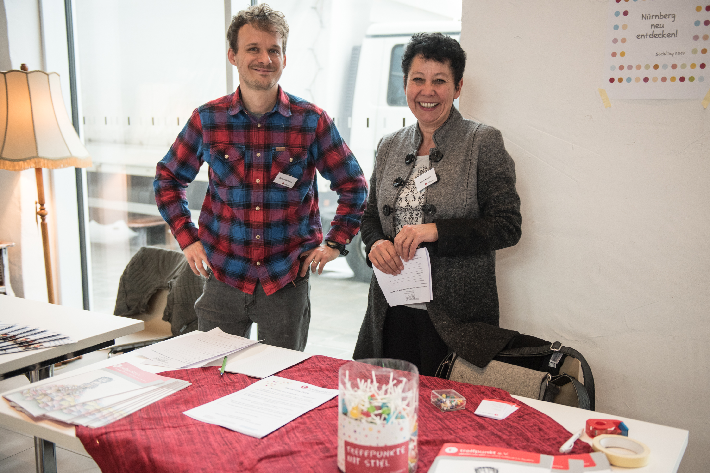 Ein junger Mann mit karriertem Hemd und eine Frau mit kurzen schwarzen Haaren stehen hinter einem Tisch mit roter Tischdecke und lächeln zufrieden in die Kamera.