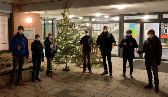 Beleuchteter Weihnachtsbaum vor Eingangstür eines Seniorenheimes bei Dunkelheit, der Baum ist flankiert von 7 Männern und Frauen in winterlicher Kleidung