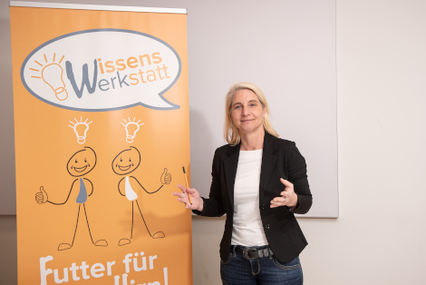 WissensWerkstatt Einre Frau steht lächelnd vor dem Rollup mit der Aufschrift "WissensWerkstatt-Futter für dein Gehirn"