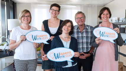 WissensWerkstatt Vier Frauen und ein Mann lächeln in die Kamera. Sie halten Sprechblasen mit dem eingedruckten Text "Wissenswerkstatt" in den Händen.
