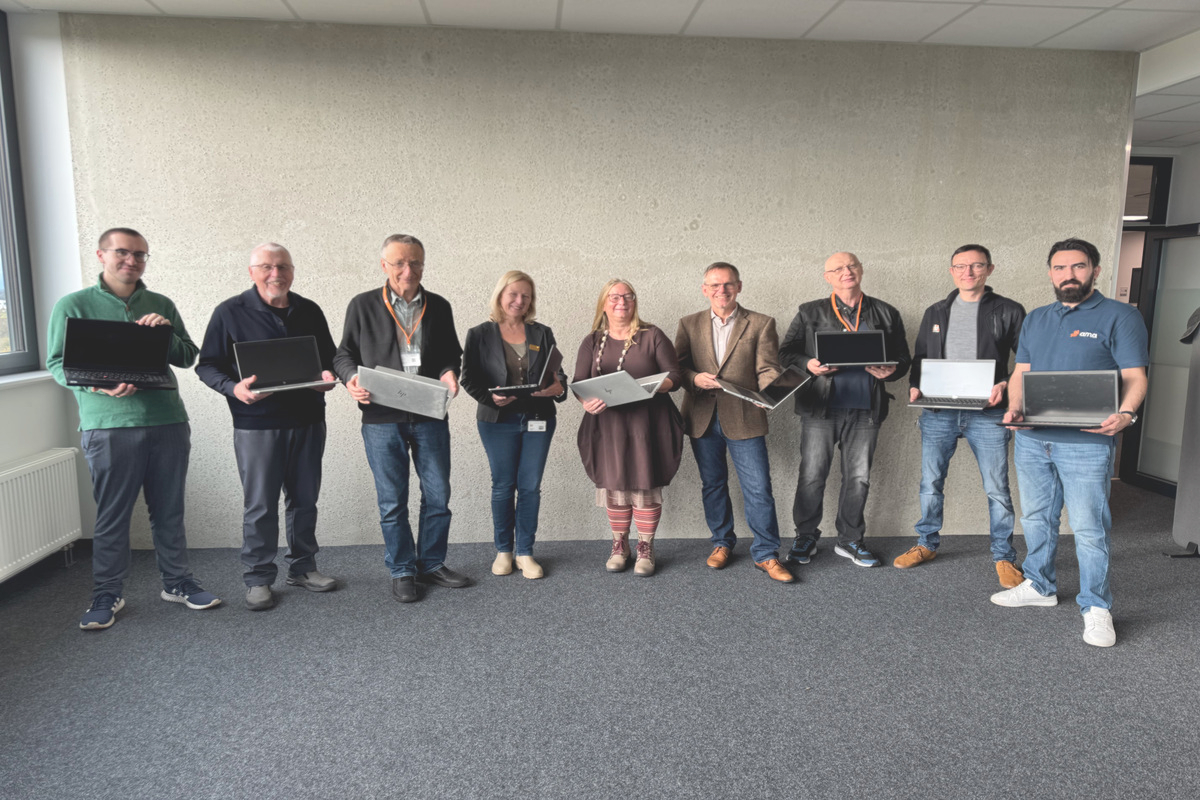 Neun Personen stehen vor einer Wand, jede Person hat einen aufgeklappten Laptop in der Hand.