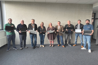 Neun Personen stehen vor einer Wand, jede Person hat einen aufgeklappten Laptop in der Hand.