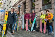 Nürnbergs Umweltreferentin Britta Walthelm (re.) und sieben Teilnehmende am Inklusiven Freiwilligentag stehen fröhlich lachend hinter ihren Kunstwerken, 20 Holzstehlen mit fröhlichen bunten Gesichtern und einem Mundloch für den Aschenbecher